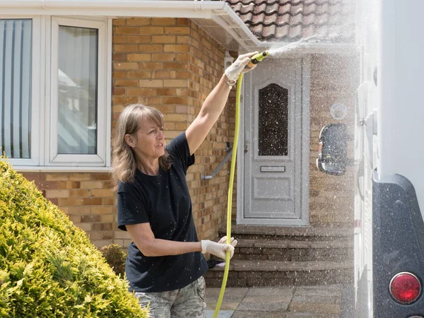 A lady motorhome owner stands cleaning her recreational vehicle with a hose pipe.Spray can be seen from the pipe