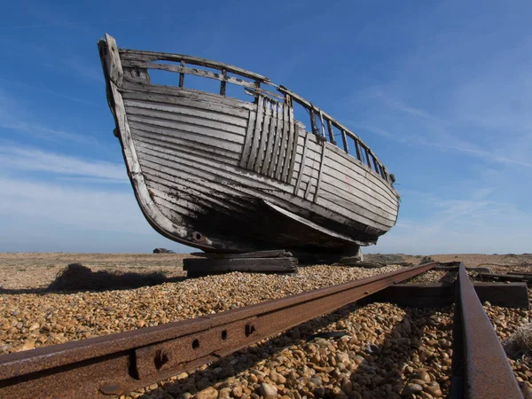 An old abandoned fishing boat sits on a shingle beach with fishing nets in foreground.A rusty disused rail line is in foreground and leads to distance