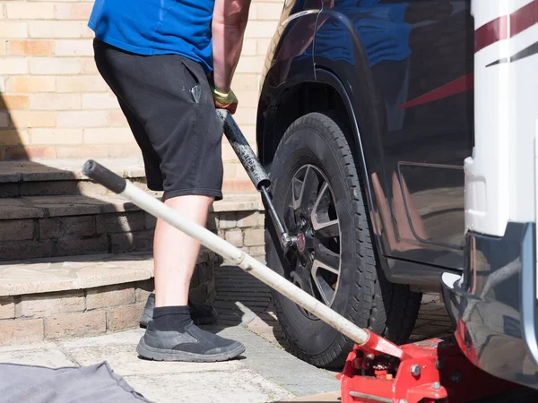 A mechanic uses a metal pole on a wrench to remove the wheel nuts from a motorhome wheel on a private drive.Trolley jack visible blurred in foreground.Man wears shorts