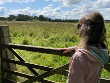 Pembe kapüşonlu bir kadın, güneş gözlüğü takarak arka plandaki vahşi bir çayıra doğru eğiliyor. Mavi gökyüzü ve beyaz bulutlarla güneşli bir gün.
