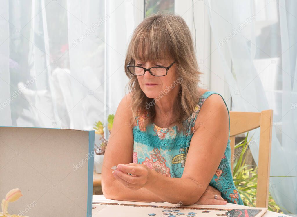 An attractive lady wearing glasses concentrates hard looking at the box lid of a jigsaw choosing where to place a jigsaw piece which she has in her hand.Jigsaw puzzle.In conservatory with net drapes in background.