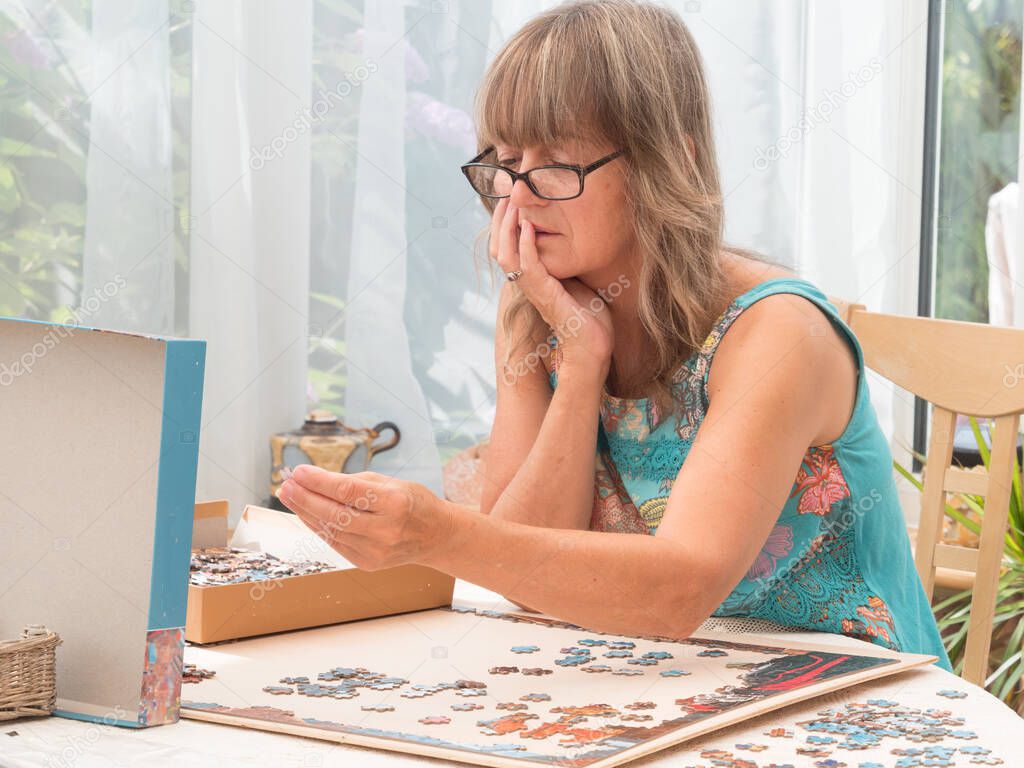 An attractive lady wearing glasses concentrates hard with chin in hand looking at the box lid of a jigsaw choosing where to place a jigsaw piece which she holds in her hand.Jigsaw puzzle.In conservatory with white net drapes in background.