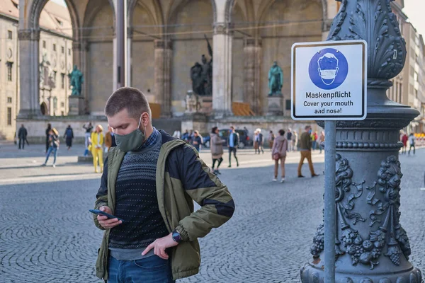 Man in a protective face mask near the sign that warns people to cover their mouth and nose at Odeon square in Munich, Germany. Man wearing a mask is holding his phone at Odeonplatz