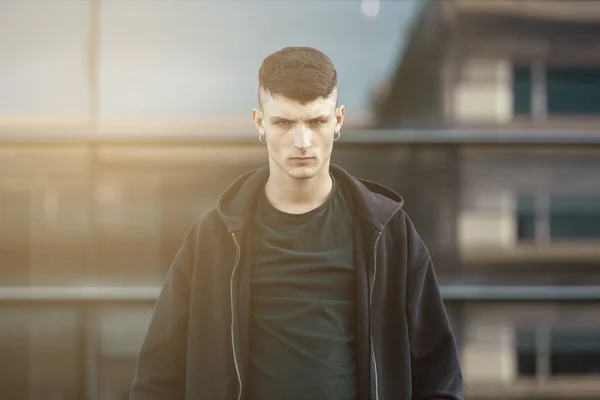 Portrait of a young man with hoop earrings and black clothes, serious expression, looking at the camera on a office building background. Young and teenage job concept