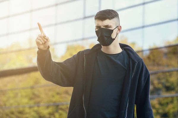Portrait of a young man, wearing a mask and black clothes, pointing upwards on a office building background. Young and teen coronavirus concept
