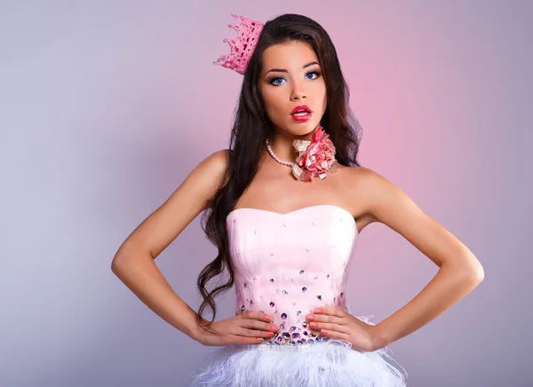 Menina morena alegre bonita em um vestido rosa e coroa rosa em sua cabeça. mini com lantejoulas e penas. fundo cinza . — Fotografia de Stock