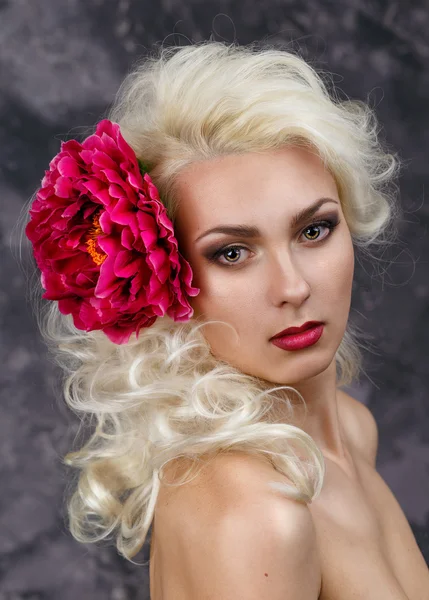 Beauty portrait of a blonde with a big red flower in her hair on a gray background. — Stock Photo, Image