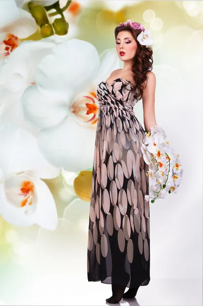 Fantasy girl in long dress with flowers in her hair and with a bouquet of orchids in their hands. — Stock Photo, Image