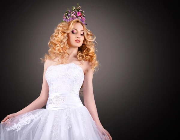 Die Braut in einem herrlichen weißen Hochzeitskleid vor grauem Hintergrund. Schönheitsporträt einer Blondine mit perfektem Make-up und einer Blütenkrone auf dem Kopf. — Stockfoto
