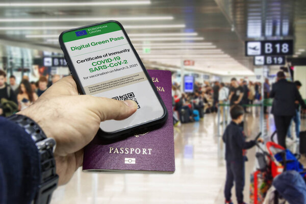 Male hand holding a phone with the European Union vaccination certificate on the screen and a traditional passport inside an airport. Immunity from Covid-19. Travel without restrictions.