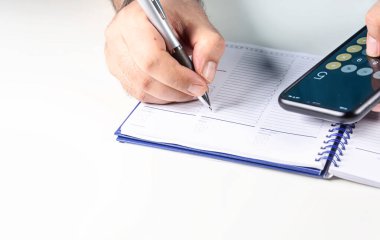 Adam cep telefonu hesap makinesiyle hesaplamalar yapıyor ve bir kalem ile klasör defterine yazıyor. Bütçe ve ekonomi.