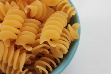 Beyaz arka planda mavi kasede çiğ makarna fusili. Mutfak masasında sağlıklı besinler var. Genel görünüm, boşluğu kopyala