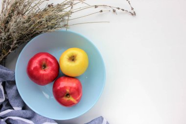 Kırmızı ve yeşil elma mavi kasede. Düz kompozisyon, taze meyveler, kuru lavanta çiçekleri ve beyaz masa arkasında çizgili gri peçete. Boşluğu kopyala, genel görünüm