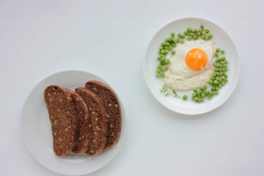 Beyaz tabakta yeşil bezelyeli sahanda yumurta ve beyaz masa arkasında çavdar ekmeği. Sağlıklı kahvaltı ya da brunch konsepti. Sunny Side ve sebzeler bir tabakta servis edilir. Üst görünüm.