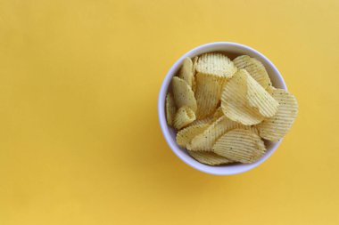 Sarı kağıtta beyaz bir kasede patates cipsi. Üst görünüm, boşluğu kopyala. Düz zemin üzerinde yemek yatıyordu