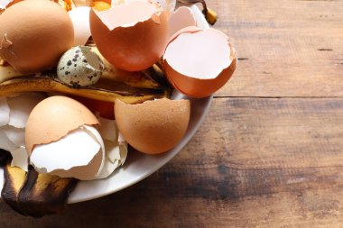 Muz derisi, yumurta kabuğu ve ahşap masa arkasında beyaz kasede portakal kabukları. Gübrelerde ya da ev yapımı gübrelerde mutfak kırıntıları kullanmak. Sıfır atık. Üst görünüm, alanı kopyala