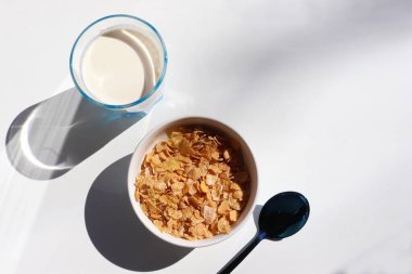 Beyaz arka planda bir kase mısır gevreği ve bir bardak süt. Mısır gevreği ve sütle sağlıklı bir kahvaltı. Üst görünüm, boşluğu kopyala. Sert ışık, gölge