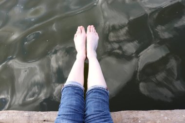 Nehir suyunun arka planında, ahşap iskelede, mavi kot giyen çıplak ayaklı bir kadın. Yaz tatili kavramı. Üst görünüm