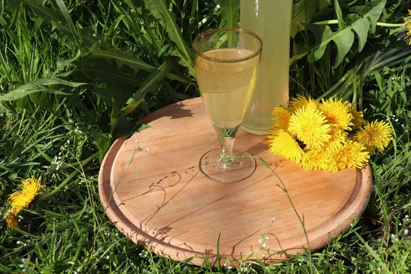 Glass of dandelion wine on wooden board in summer garden. Homemade herbal drink in bottle on green grass with yellow flowers background.