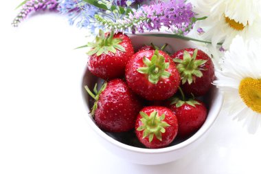 Fresh strawberries in a bowl with bouquet of summer flowers on white table. Top view, copy space. Midsummer still life