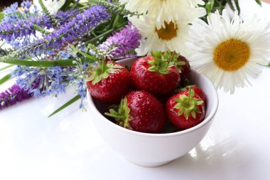 Fresh strawberries in a bowl with bouquet of summer flowers on white table. Top view, copy space. Midsummer still life