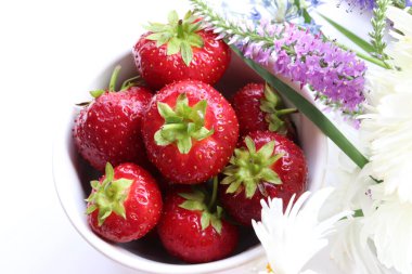 Fresh strawberries in a bowl with bouquet of summer flowers on white table. Top view, copy space. Midsummer still life