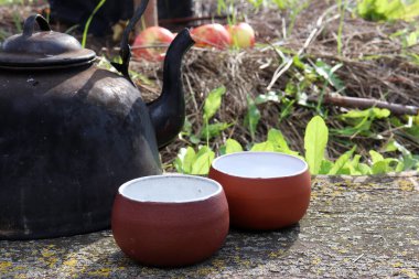 Eski siyah su ısıtıcısı ve iki el yapımı seramik bardak bahçede yeşil çimen arka planında ahşap bir masa üzerinde