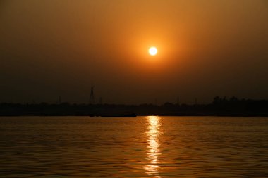 A dazzling golden sun and its reflection dominate the river scene, setting a warm, bright glow against the stark, dark profile of the distant shore