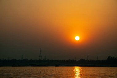 A warm, bright light from the brilliant golden sun and its mirrored reflection completely overwhelms the river, sharply with the darkness of the far shore