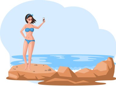 Mavi mayo ve güneş gözlüklü genç bir kadın sahilde taşın üzerinde duran akıllı telefonuyla selfie çekiyor.