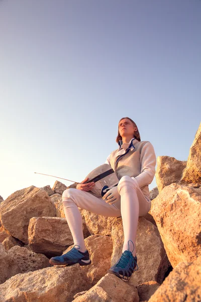 Fencer man sitting on top of the rock holding his fencing mask and a sword on a blue sky background and looking forward dreamingly — Stock Photo, Image