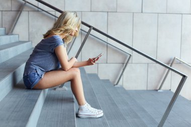 Girl typing message on mobile phone and sitting on stairs clipart