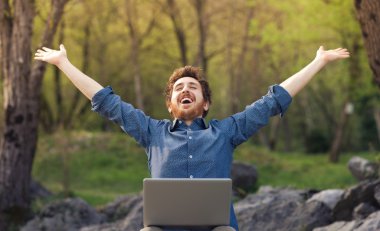 Happy hipster with laptop in the forest clipart