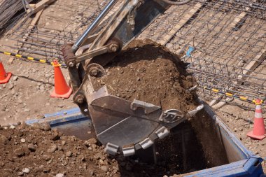 Kazı kovası kamyon yatağına ağır miktarda toprak boşaltıyor. Bu da inşaat alanındaki ilerlemenin sabit ritmine güç veriyor..