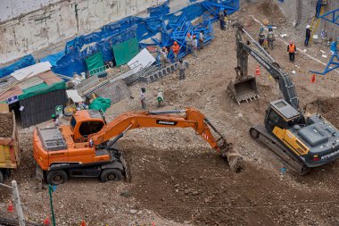 İki kazıcı, dinamik kazı ortamında işçiler, güvenlik ekipmanları ve yapısal malzemelerle çevrili bir inşaat sahasında aktif toprak taşımacılığı yapıyor..