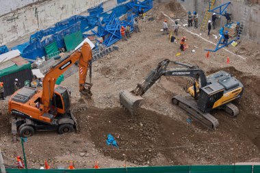 İki kazıcı, dinamik kazı ortamında işçiler, güvenlik ekipmanları ve yapısal malzemelerle çevrili bir inşaat sahasında aktif toprak taşımacılığı yapıyor..