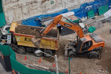 İki kazıcı, dinamik kazı ortamında işçiler, güvenlik ekipmanları ve yapısal malzemelerle çevrili bir inşaat sahasında aktif toprak taşımacılığı yapıyor..