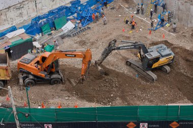 İki kazıcı, dinamik kazı ortamında işçiler, güvenlik ekipmanları ve yapısal malzemelerle çevrili bir inşaat sahasında aktif toprak taşımacılığı yapıyor..