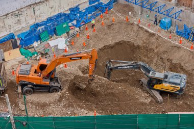 İki kazıcı, dinamik kazı ortamında işçiler, güvenlik ekipmanları ve yapısal malzemelerle çevrili bir inşaat sahasında aktif toprak taşımacılığı yapıyor..