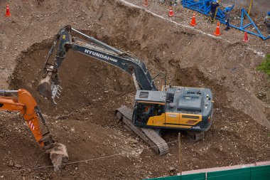 İki kazıcı, dinamik kazı ortamında işçiler, güvenlik ekipmanları ve yapısal malzemelerle çevrili bir inşaat sahasında aktif toprak taşımacılığı yapıyor..