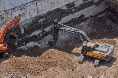 İki kazıcı, dinamik kazı ortamında işçiler, güvenlik ekipmanları ve yapısal malzemelerle çevrili bir inşaat sahasında aktif toprak taşımacılığı yapıyor..
