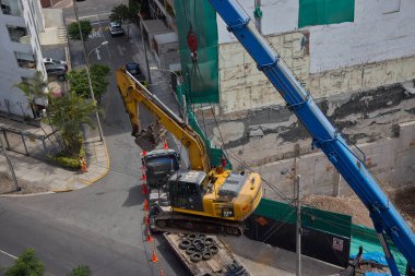 Ağır vinç kazıcıyı şehir ortamında derin bir inşaat sahasına kaldırıyor, altyapı geliştirme, gayrimenkul büyümesi ve modern şehir genişlemesi sergiliyor..