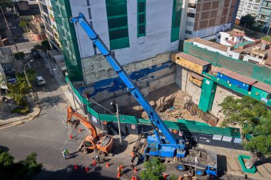 Ağır vinç kazıcıyı şehir ortamında derin bir inşaat sahasına kaldırıyor, altyapı geliştirme, gayrimenkul büyümesi ve modern şehir genişlemesi sergiliyor..