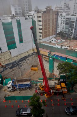 Bir vinç Miraflores, Lima 'daki kentsel bir inşaat sahasında, temel çalışması sırasında vinç operasyonları, güvenlik bariyerleri ve kontrollü lojistik faaliyetlerini gösteren bir inşaat malzemesi çıkarıyor. Sahne profesyonelliği vurguluyor.