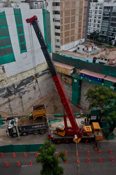 Bir vinç Miraflores, Lima 'daki kentsel bir inşaat sahasında, temel çalışması sırasında vinç operasyonları, güvenlik bariyerleri ve kontrollü lojistik faaliyetlerini gösteren bir inşaat malzemesi çıkarıyor. Sahne profesyonelliği vurguluyor.