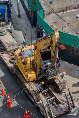 Bir vinç derin bir kazı çukuru içindeki kazıcıyı tam olarak kaldırdığı, sıkı güvenlik protokolleri, koordine takım çalışması ve ağır kentsel mühendislik operasyonlarında yüksek çalışma standartları gösteren bir inşaat alanının yüksek açılı görüntüsü..