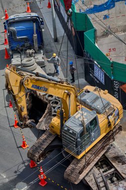Bir vinç derin bir kazı çukuru içindeki kazıcıyı tam olarak kaldırdığı, sıkı güvenlik protokolleri, koordine takım çalışması ve ağır kentsel mühendislik operasyonlarında yüksek çalışma standartları gösteren bir inşaat alanının yüksek açılı görüntüsü..