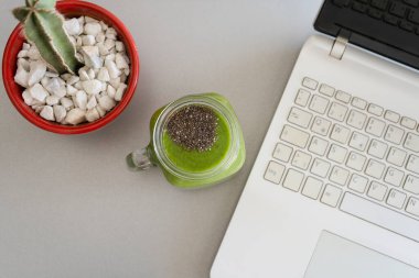 Gri arka planda yeşil kivi smoothie, laptop ve kaktüs içeren sağlıklı bir işyeri. Evde çalış, mola ver, serbest çalış ve dijital göçebe kavramı.