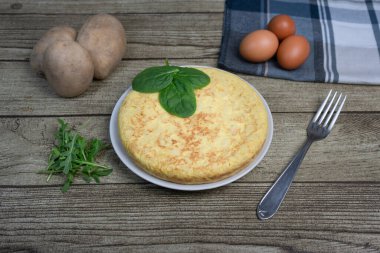 Tabakta Tortilla de patatas adı verilen İspanyol patates omleti, ahşap arka planda taze yapraklar, yumurtalar ve patatesler..