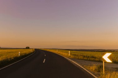 Gün batımında kırsal alanda trafik işaretleri ve tarlaları olan şaşırtıcı asfalt kıvrımlı bir yol. Güneşli bir gün..